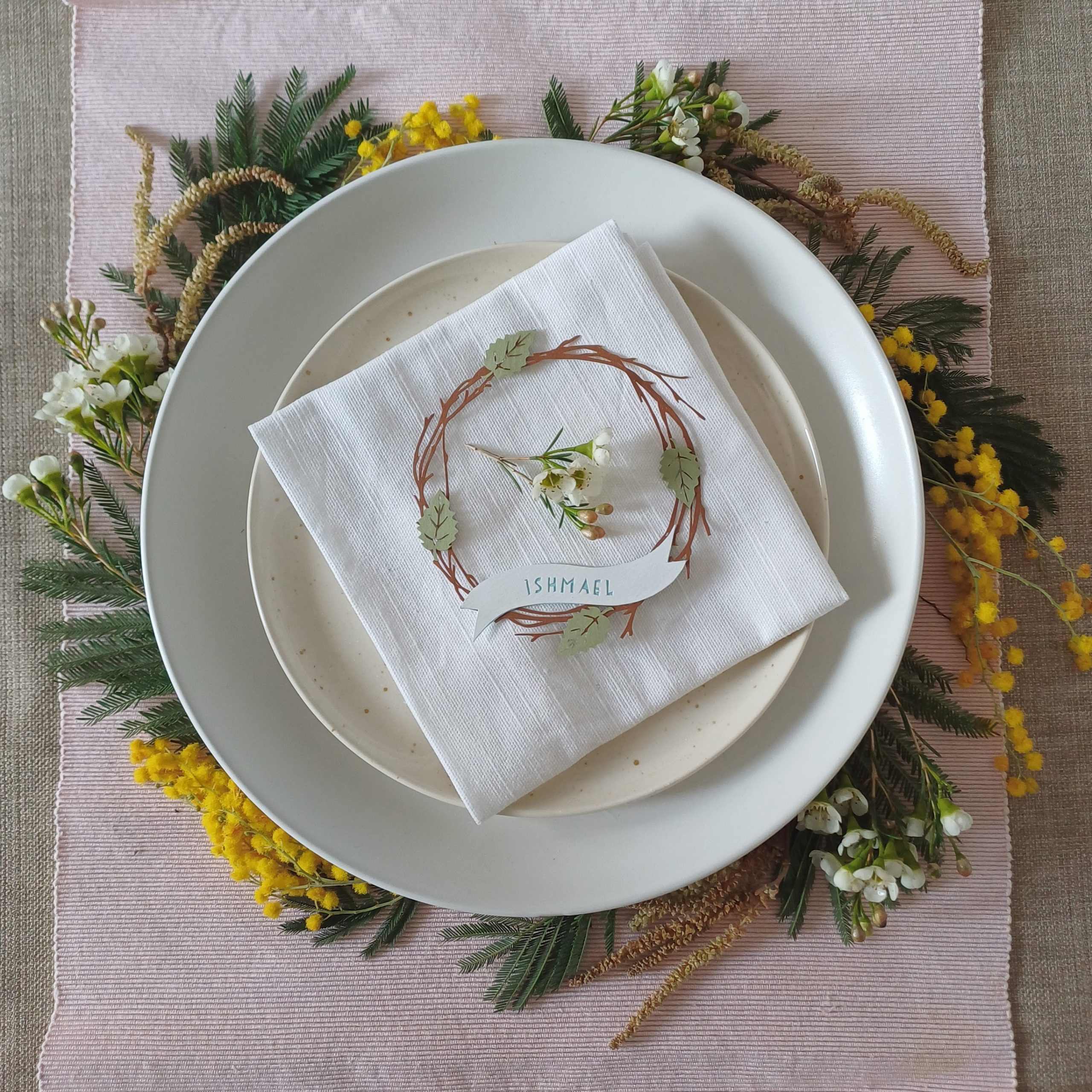 Lasercut birch twig wreath made from dark brown art paper, with 4 pale green paper leaves distributed evenly around the wreath. At the bottom of the wreath is a white paper banner with a name Sophie cut out to reveal the letters in yellow. Place settings is placed on a napkin, on top of stacked plates which sit on a circle of spring flowers and foliage.