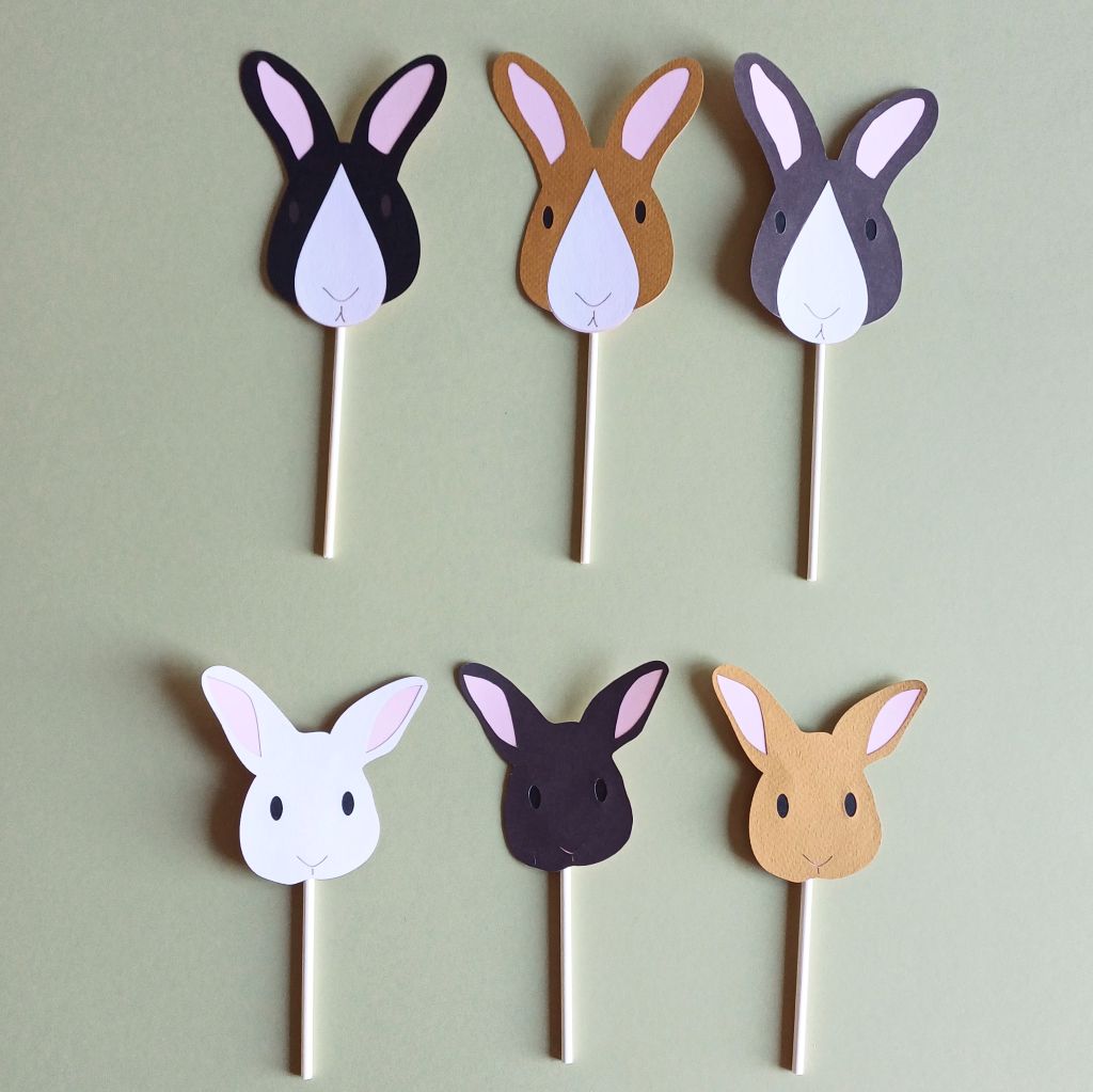 Brown, black and white bunny cake topper option. Top row: 3 bunny cake toppers (left to right) Black rabbit with dark brown eyes, pale pink ears and a white nose patch, toffee brown bunny with black eyes, pale pink ears, white nose patch, dark grey bunny with pale pink ears, black eyes, white nose patch. Bottom row (left to right) white, dark brown and toffee brown bunnies all with black eyes and pale pink ears. Each cake topper is attached to a white paper cake-pop stick