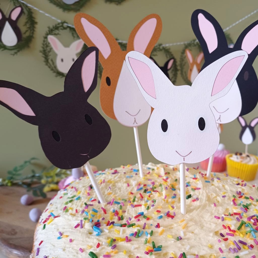 Closeup view of Dark brown and White Bunny faces with pale pink ears, small black eyes, line detail mouth and nose. In the background co-ordinating Easter Bunny Papercut Garland is hung on a sage green wall.