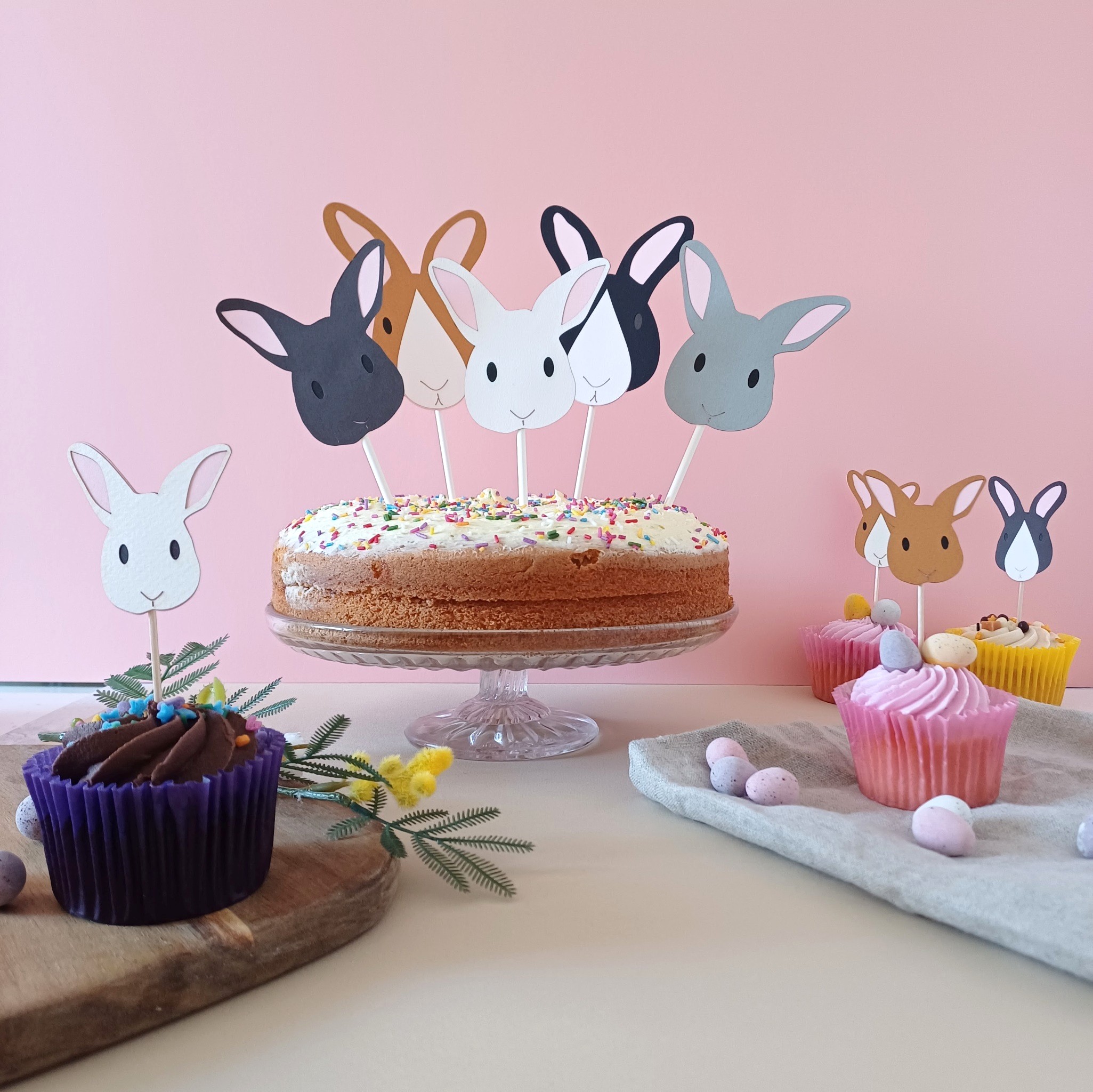 Easter bunny cake toppers displayed in a cake on a cake stand. Easter bunny cupcakes are placed on the table around the large cake.