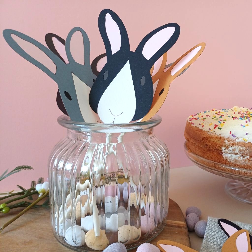 Easter bunny cake toppers placed in a ribbed glass jar filled with mini eggs. Decorations include black and white rabbit, toffee brown and white rabbit, dark grey and white rabbit.