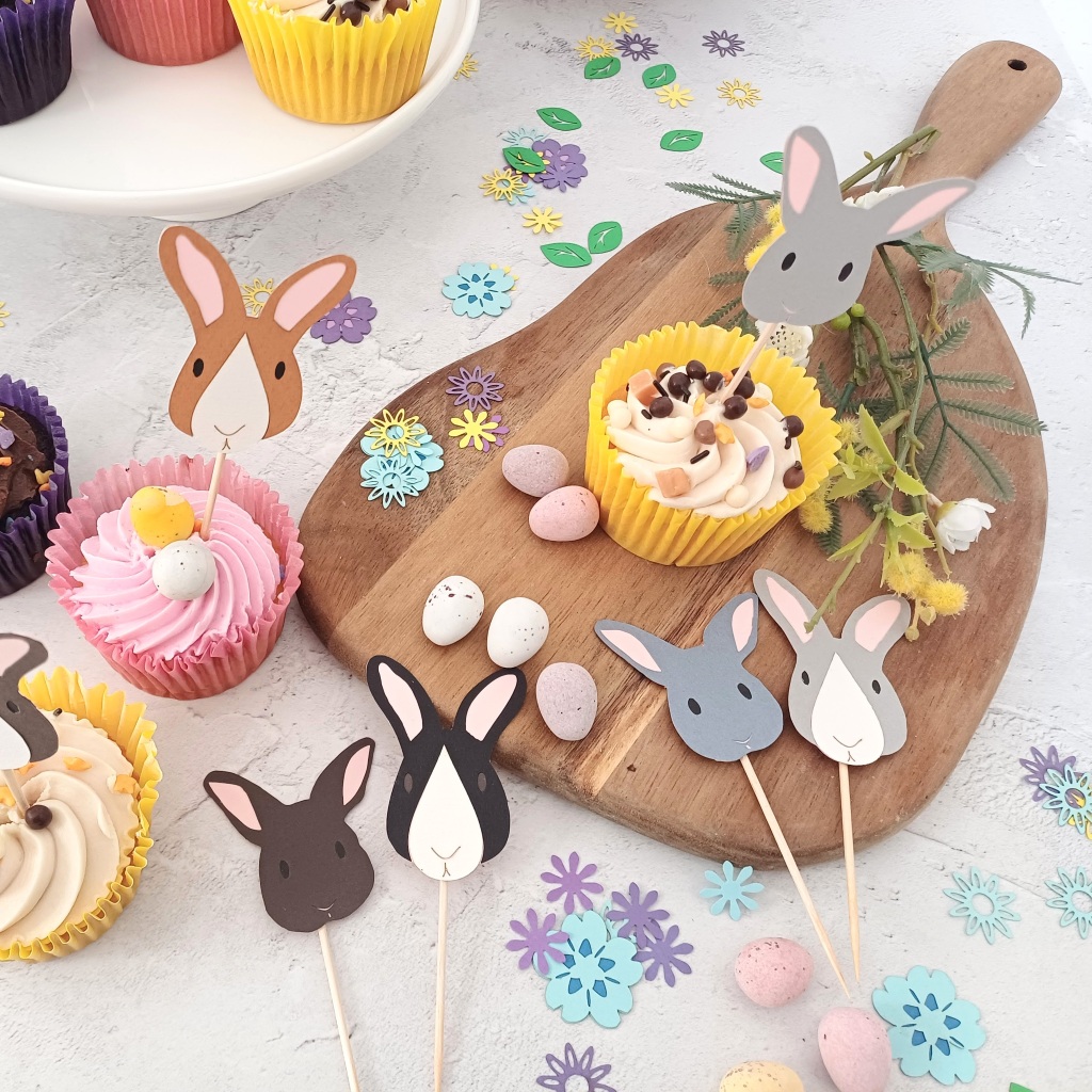 Easter Bunny Cupcake Toppers displayed on a kitchen counter. Some cake toppers are placed into iced cupcakes. Some bunny cupcake decorations lay flat on the counter. Countertop is decorated with mini eggs and paper flower confetti.