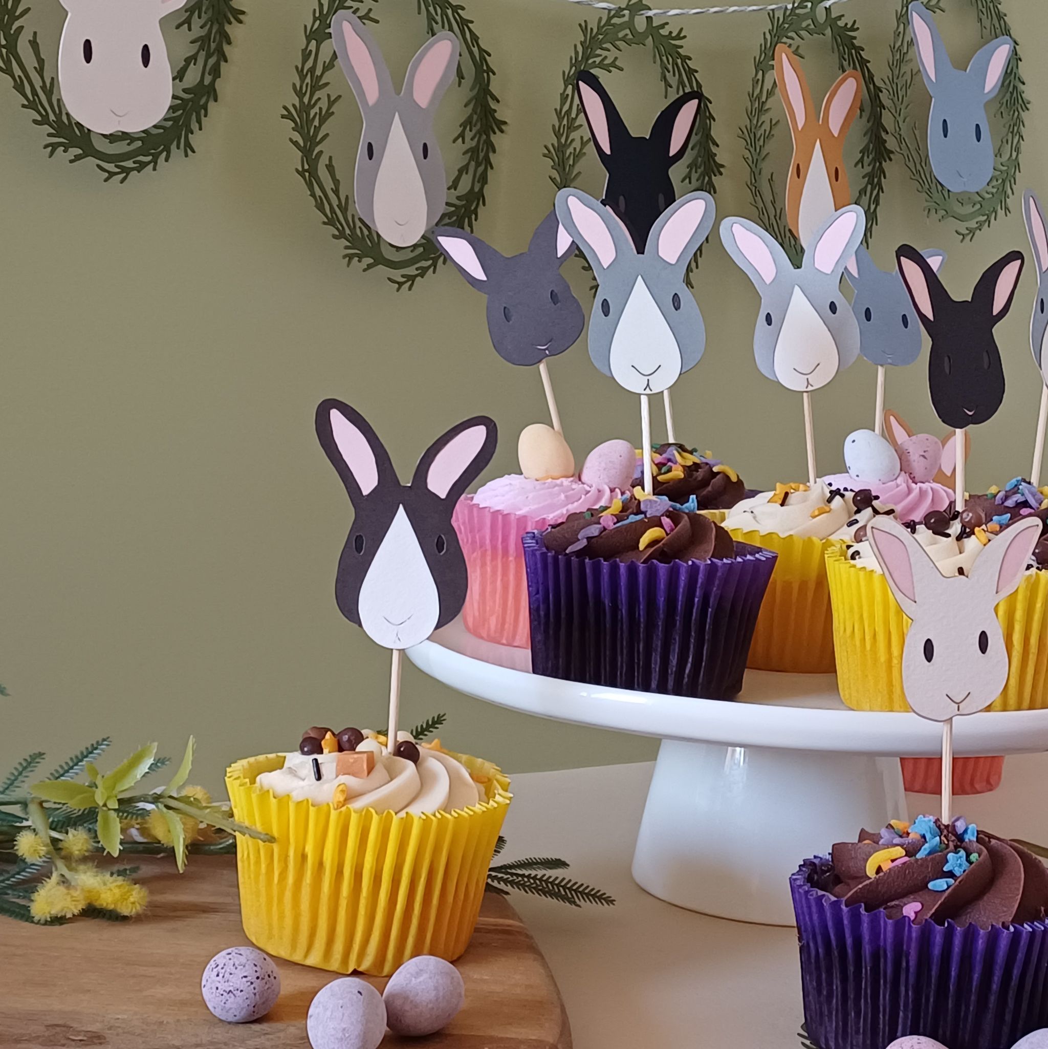Easter Bunny Cupcake Toppers Set of 12 displayed in cupcakes on cake stand and table decorated for Easter. In the background on a green wall, a coordinating Easter Bunny Garland is displayed. All bunny faces are either brown, grey, black or white.