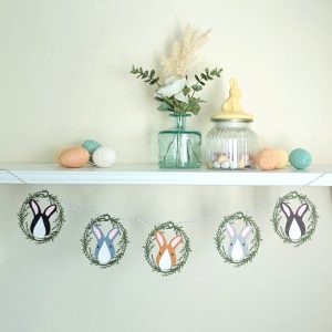 Easter garland hung from a shelf decorated with a vase of flowers and easter treats. Garland features five decorations threadedonto pale greay and white bakers twine. Each decoration is made of a lasercut card olive green foliage wreath encircling a paper easter bunny face. Bunnies all have white nose patches, pale pink ears and small oval eyes, with line details for their mouth and nose. Colours of rabbits from left to right: Black and white, Dark grey and white, Toffee brown and white, pale grey and white, dark brown and white.