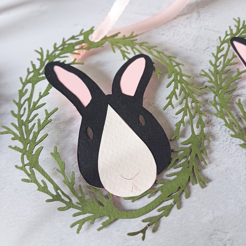 Closeup of easter tree bunny hanging decoration. Bunny face is black with a white tear drop shaped nose patch. Bunny has pale pink inner ears, small oval dark brown eyes and line details for mouth and nose. Rabbit is encircled by an oval frame of lasercut olive green foliage based on fronds of conifer.