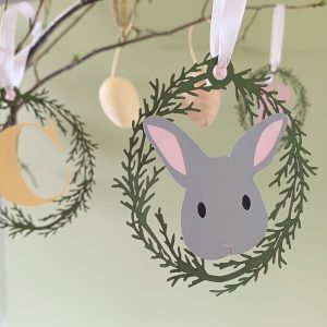 Closeup view of Easter Bunny Papercut Hanging Decoration. Decoration is hung from a branch with coordinating decorations in the backgroun. Bunny decoration features an oval of olive green lasercut foliage circling a grey bunny face. THe bunny has black eyes, pale pink ears and line details for its mouth and nose