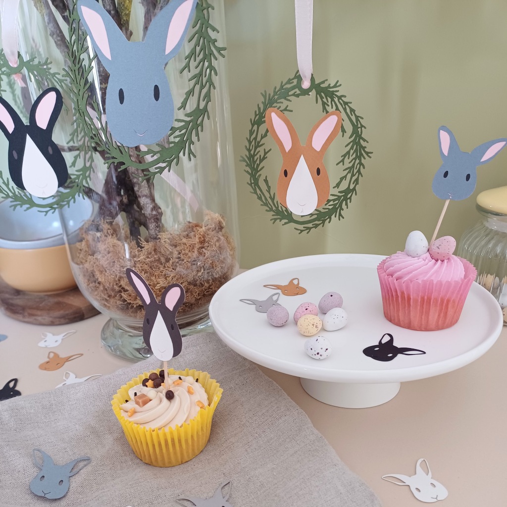 Easter Bunny Tree decorations hung from branches placed in a vase. At the foot of the vase are matching easter bunny cupcake toppers placed into iced cupcakes