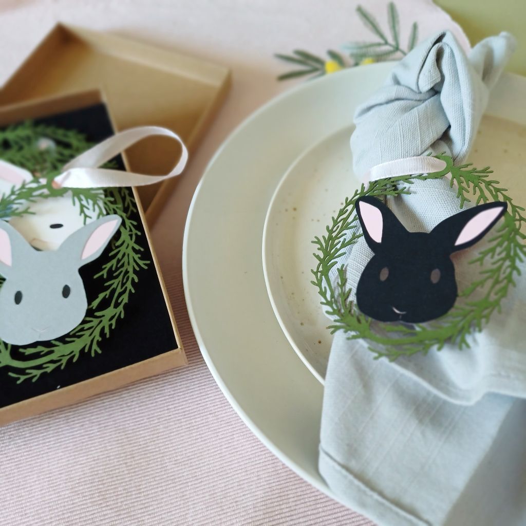 Easter Tree Bunny Hanging Decorations in cardboard presentation box on a table. These decorations are beside two stacked plates. On the top plate is a knotted linen napkin decorated with a balck bunny papercut hanging decoration that is being used as a place setting decoration