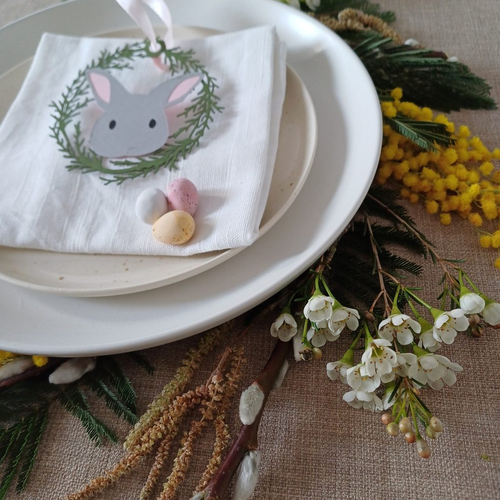 Grey Easter Bunny Place Setting Decoration sits on top of a white napkin white some mini eggs. Napkin is on top of two stacked plates which are circled with spring foliage and flowers on the tablemat underneath.