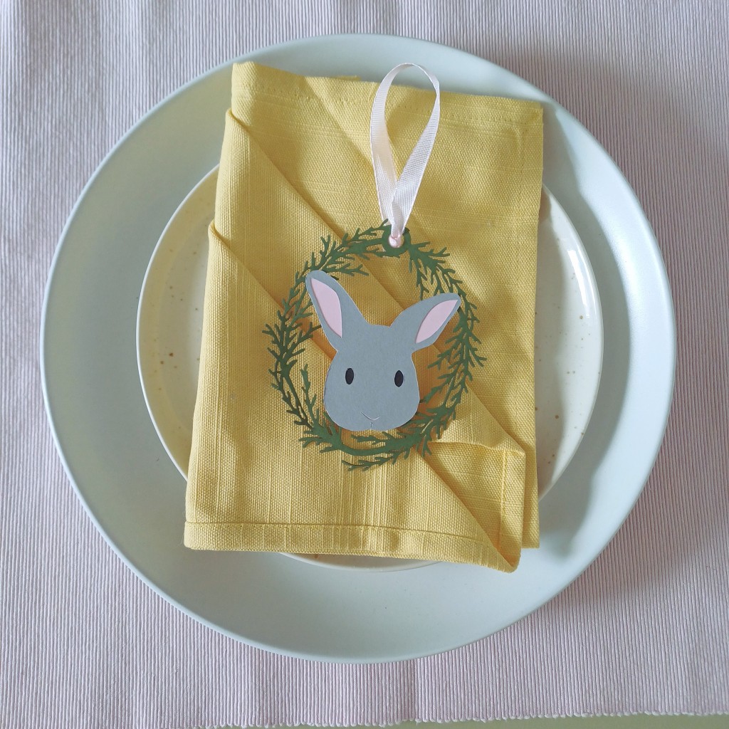 Pale Grey Easter Bunny Place Setting Decoration is shown on top of a bright yellow napkin which is folded into a rectangle with diagonal creases. Napkin sits on top of two stacked cream plates with a pink fabric tablemat underneath.