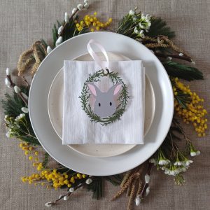 Pale Grey Easter Bunny Place Setting Decoration with pale pink ribbon loop sits in the middle of a white square napkin. Napkin is on top of two stacked cream plates. Plates are surrounded by mimosa, pussy willow, catkins and waxflower blossom circled to look like a wreath. Bunny is pale grey with smalll black oval eyes, pale pink ears and line detail for nose and mouth. Bunny is framed by olive green oval wreath of lasercut card foliage.
