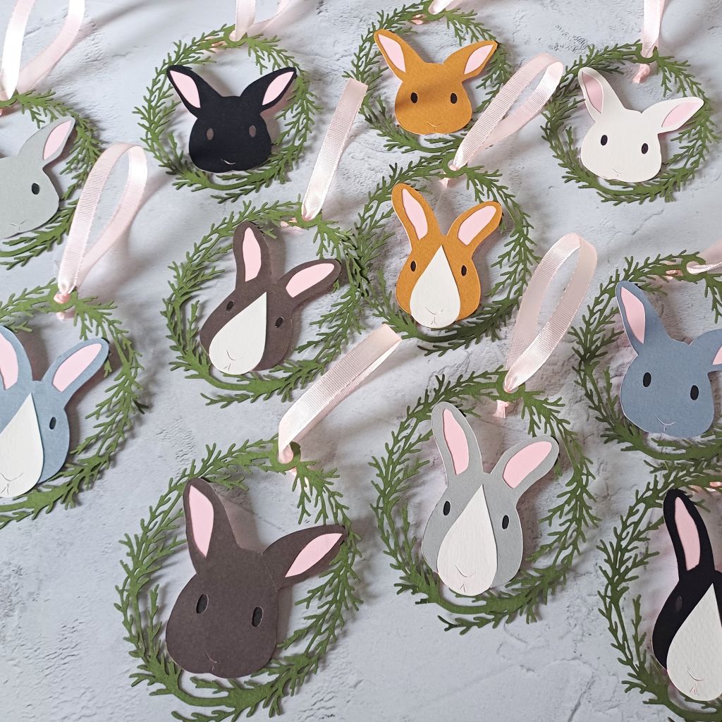 Set of 12 easter tree ornaments displayed on grey worktop. Each decoration is threaded with a pale pink grosgrain ribbon loop. Each decoration features a bunny face encircled by an oval of olive green conifer foliage frame. Rabbit faces are a mix of borwns, greys, white and black.