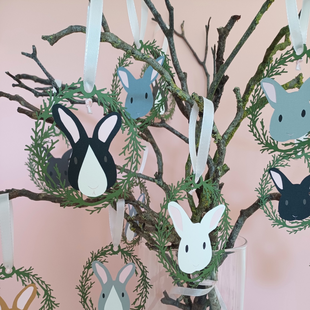 Easter tree bunny hanging decorations displayed on branches placed in a vase. Decorations include a range of bunny faces encircled by an oval wreath of green lasercut foliage. Bunny faces are a mix of greys, white and black. Decorations are threaded with pale pink grosgrain ribbon loop.