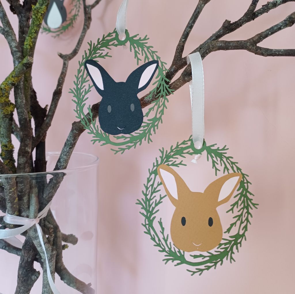 Toffee brown and black Easter tree bunny hanging decorations displayed on branches placed in a vase. Decorations include a range of bunny faces encircled by an oval wreath of green lasercut foliage. Decorations are threaded with pale pink grosgrain ribbon loop.