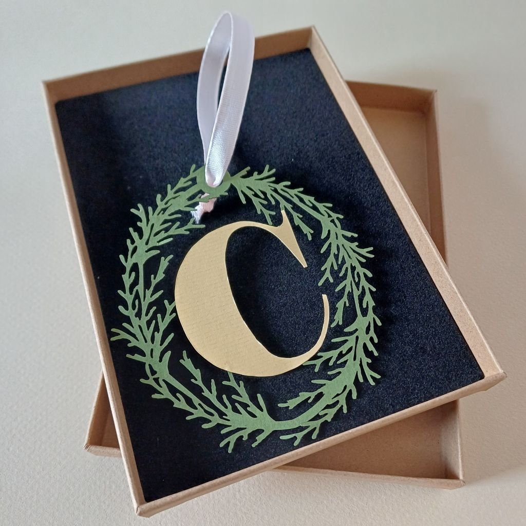 Papercut Hanging Decoration "C" in pale yellow held above cardboard presentation box with black foam insert