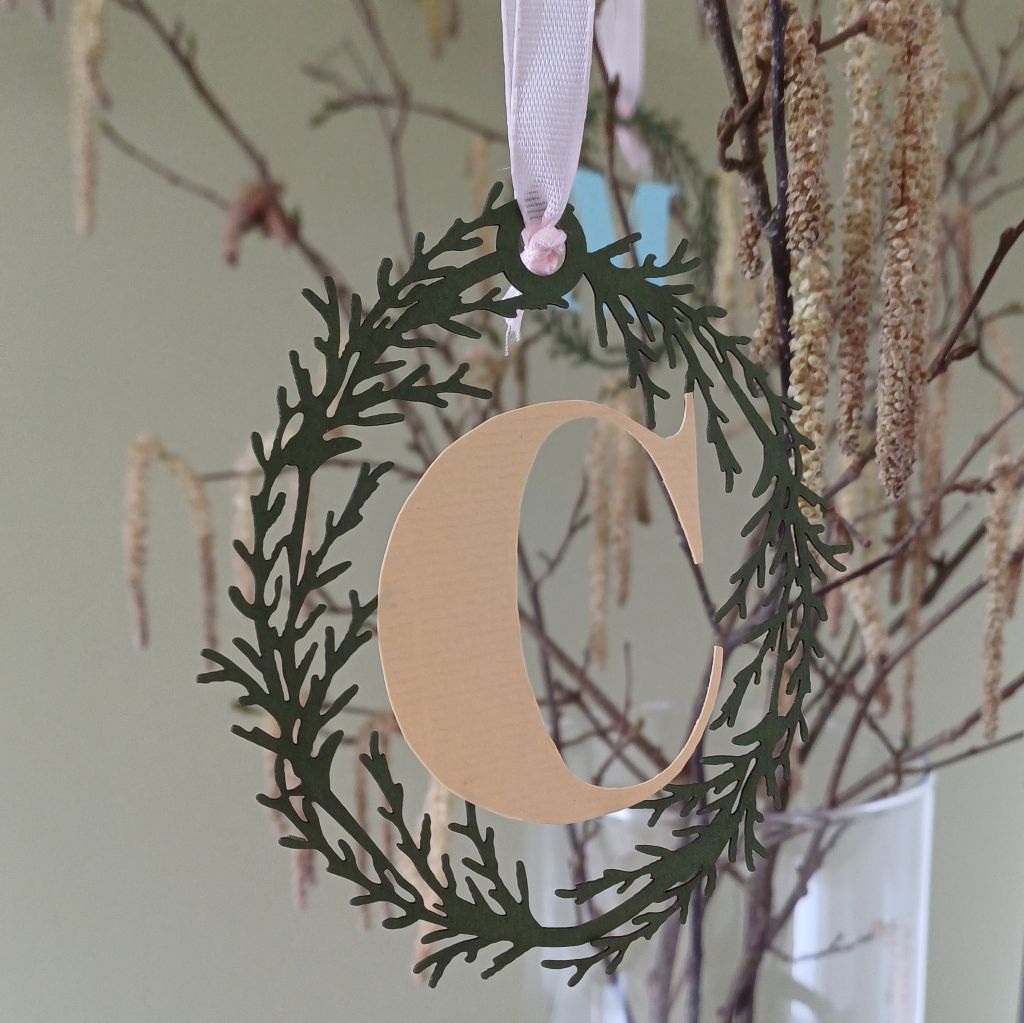 Oval Papercut Foliage wreath encircles Letter C cut in pale yellow paper. Decoratio hangs from branch with catkins