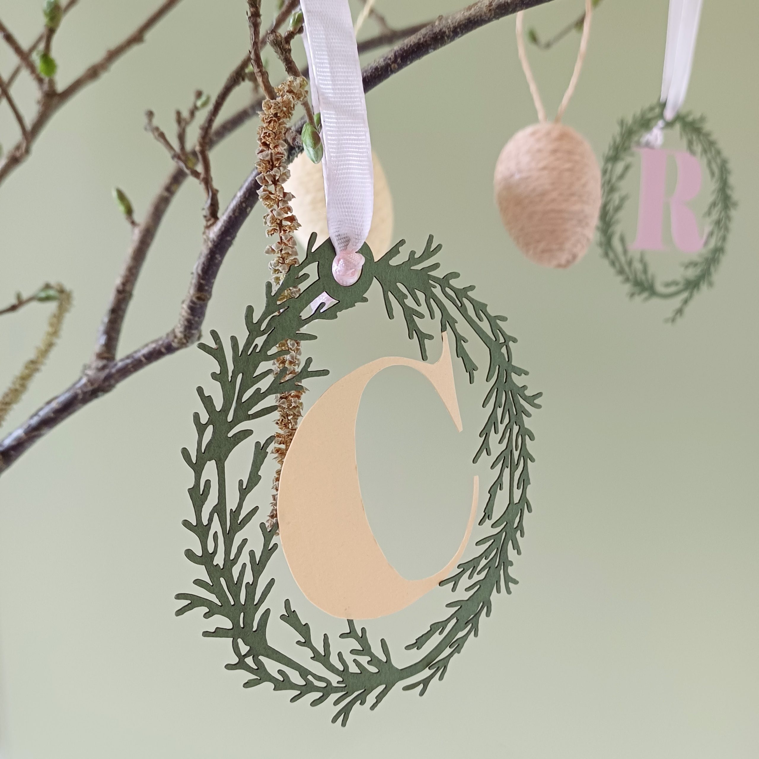 Oval Papercut Foliage wreath encircles Letter C cut in pale yellow paper. Decoration hangs from branch with catkins. Branches decorated with other paper initials and easter egg ornaments