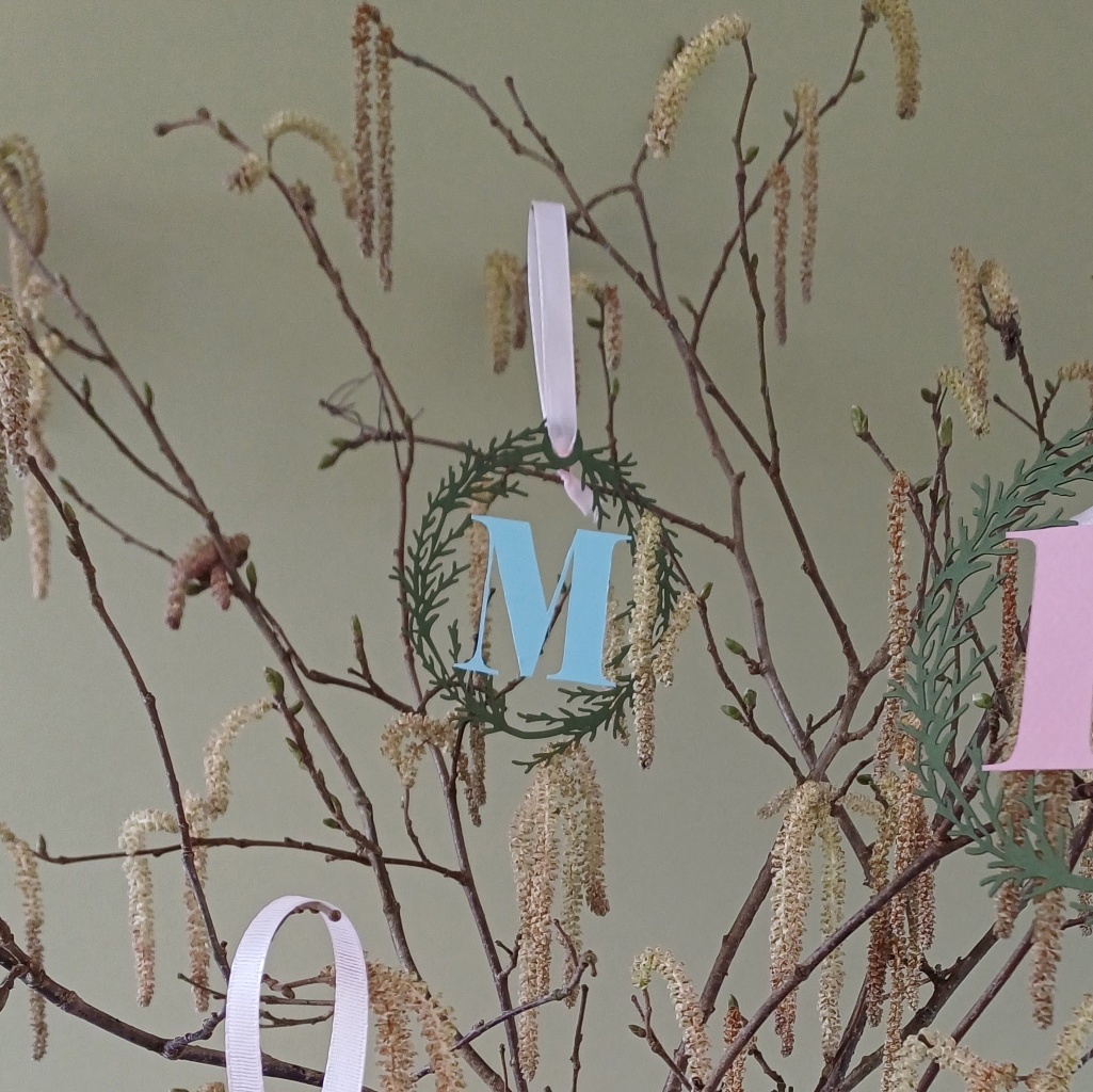 Hanging Decoration with Pale Blue Letter M surrounded by olive green foliage wreath (cut from paper) hangs on branch with spring catckins