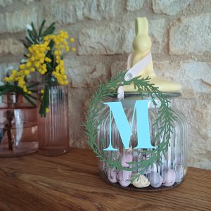 Paper Decoration made of oval papercut foliage wreath encircling plae blue letter M. Decoration hangs from Easter treat jar by pale pink ribbon hanging loop