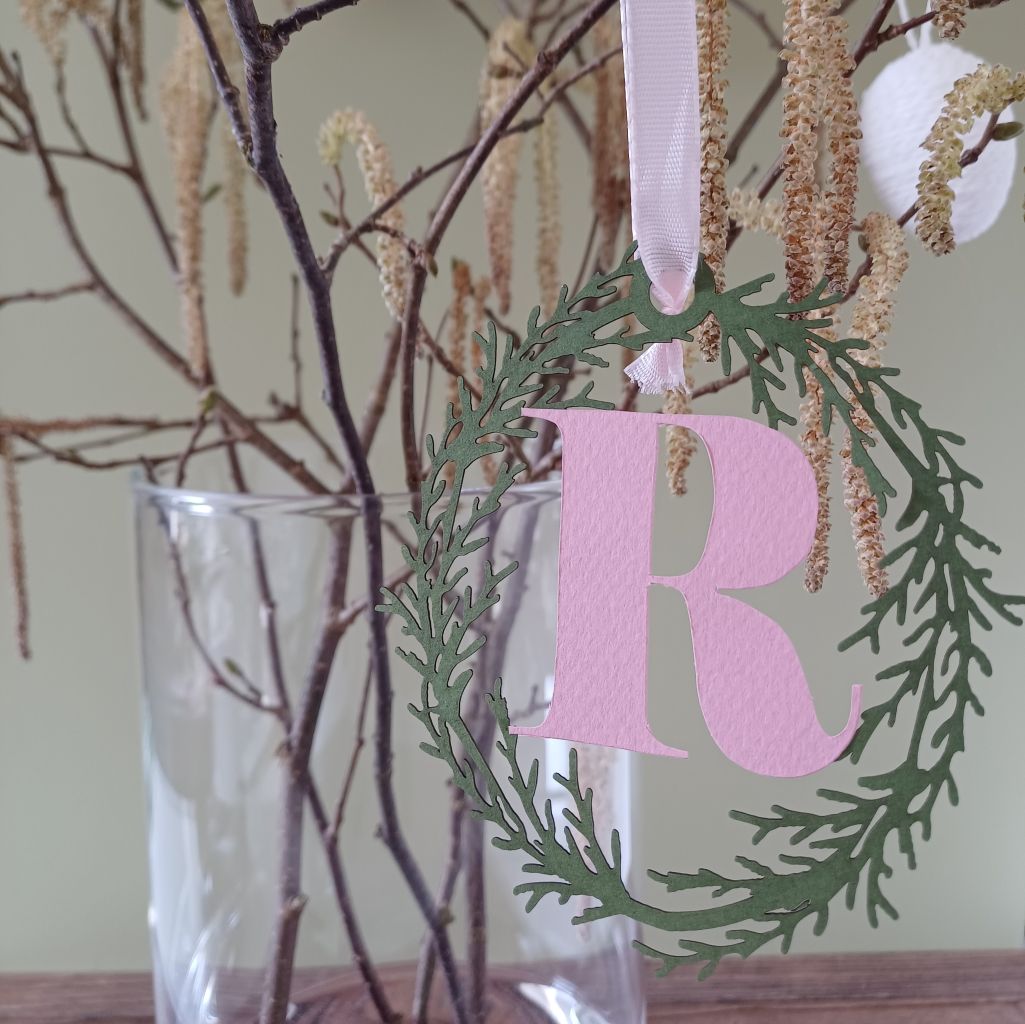 Close view of papercut Initial R in pale pink surrounded by olive green paper foliage wreath. Decoration hangs from branches placed in a vase