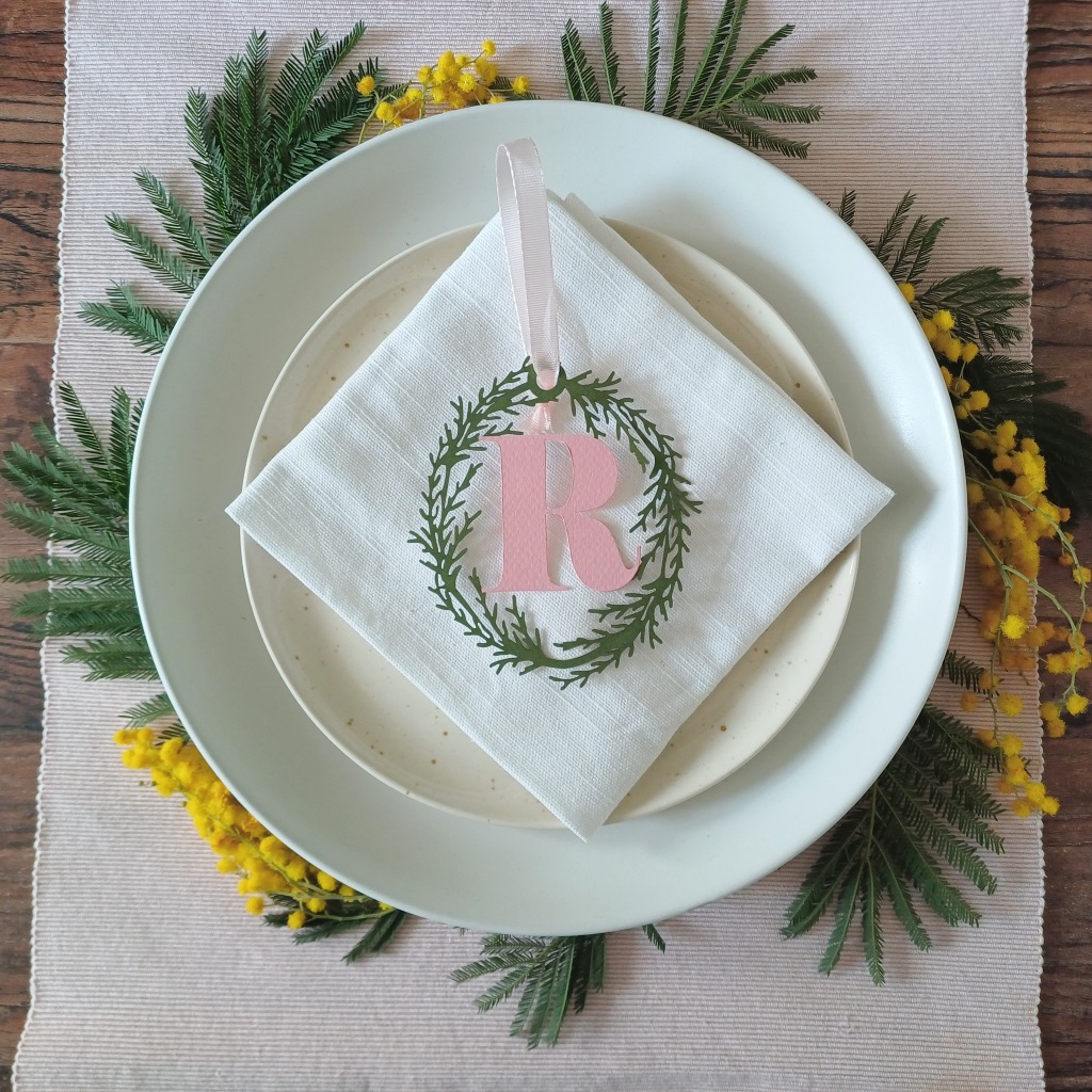 Hanging Decoration with pink initial R used as a place setting. Decoration sits on a white napkin placed on top of stacked plates surrounded by spring flowers and foliage