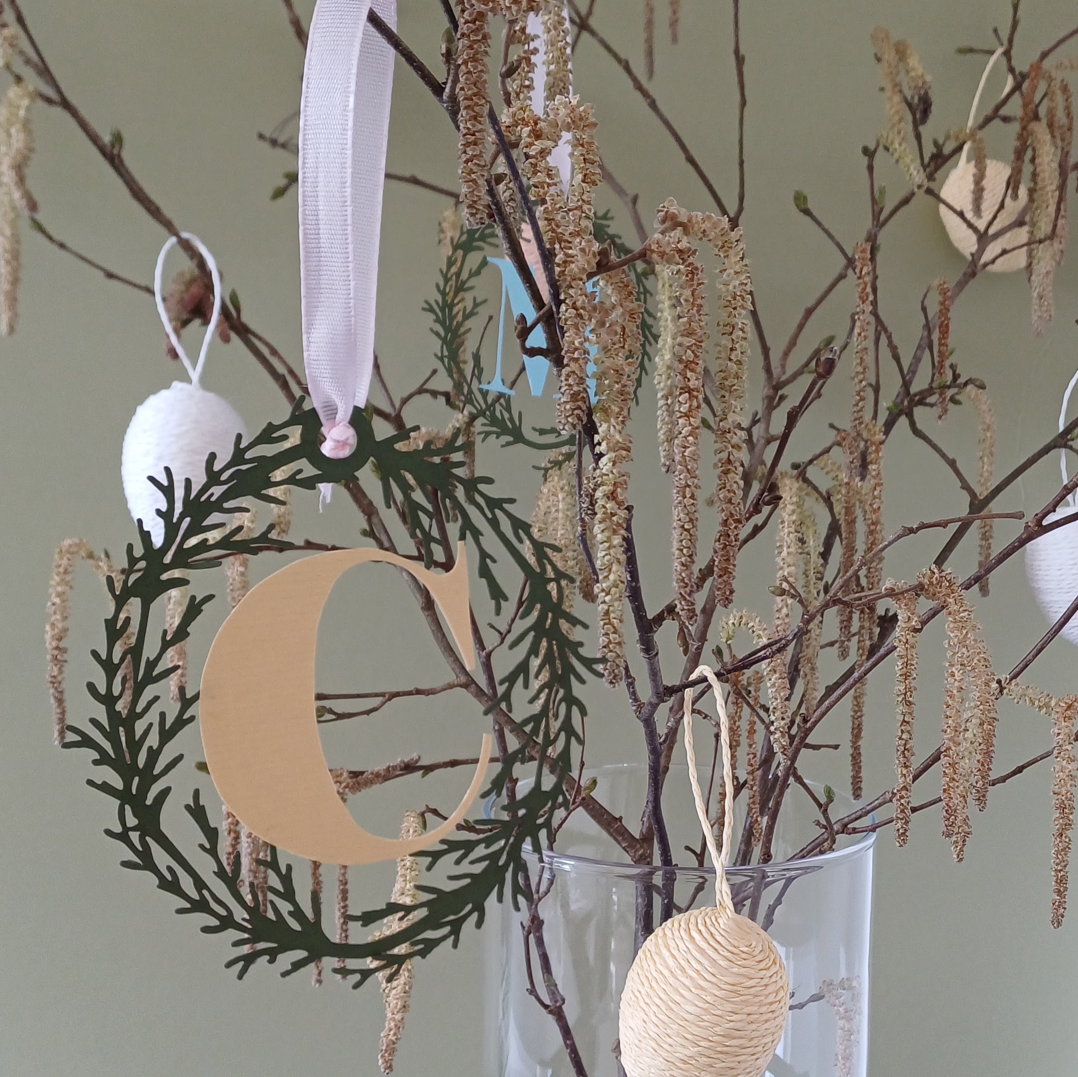 Papercut green foliage oval wreath with yellow letter C in the centre hangs by pale pink ribbon loop from catkin filled branches