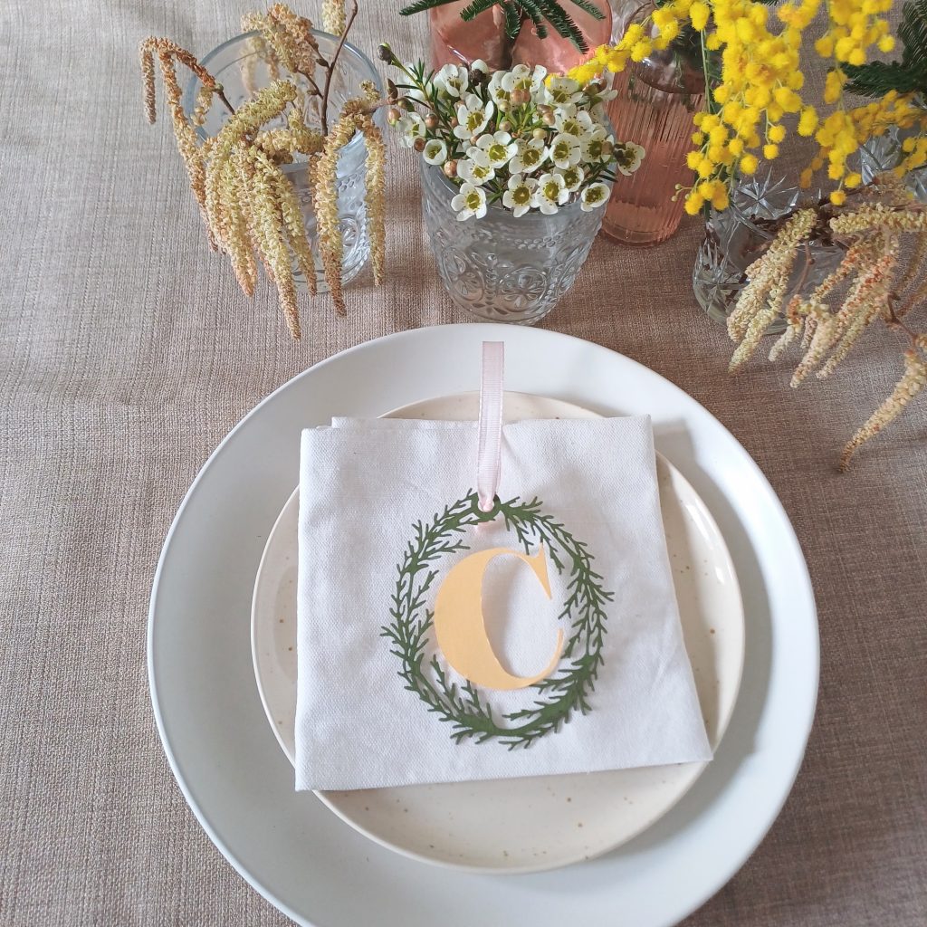 Place setting created from olive green card oval foliage wreath with papercut pale yellow letter C in the centre. Decoration is threaded with pale pink ribbon and placed on top of white napkin on cream plates