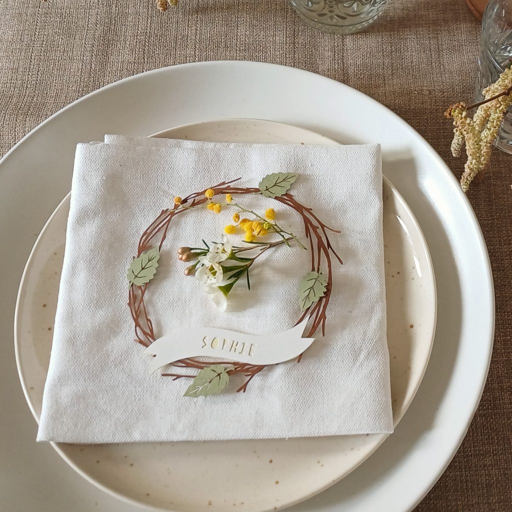 Papercut place setting made from brown paper birch wreath with 4 light green leaves. At the bottom a white banner with the name Sophie cut out in pale yellow. A sprig of mimosa and waxflower sits in the centre.