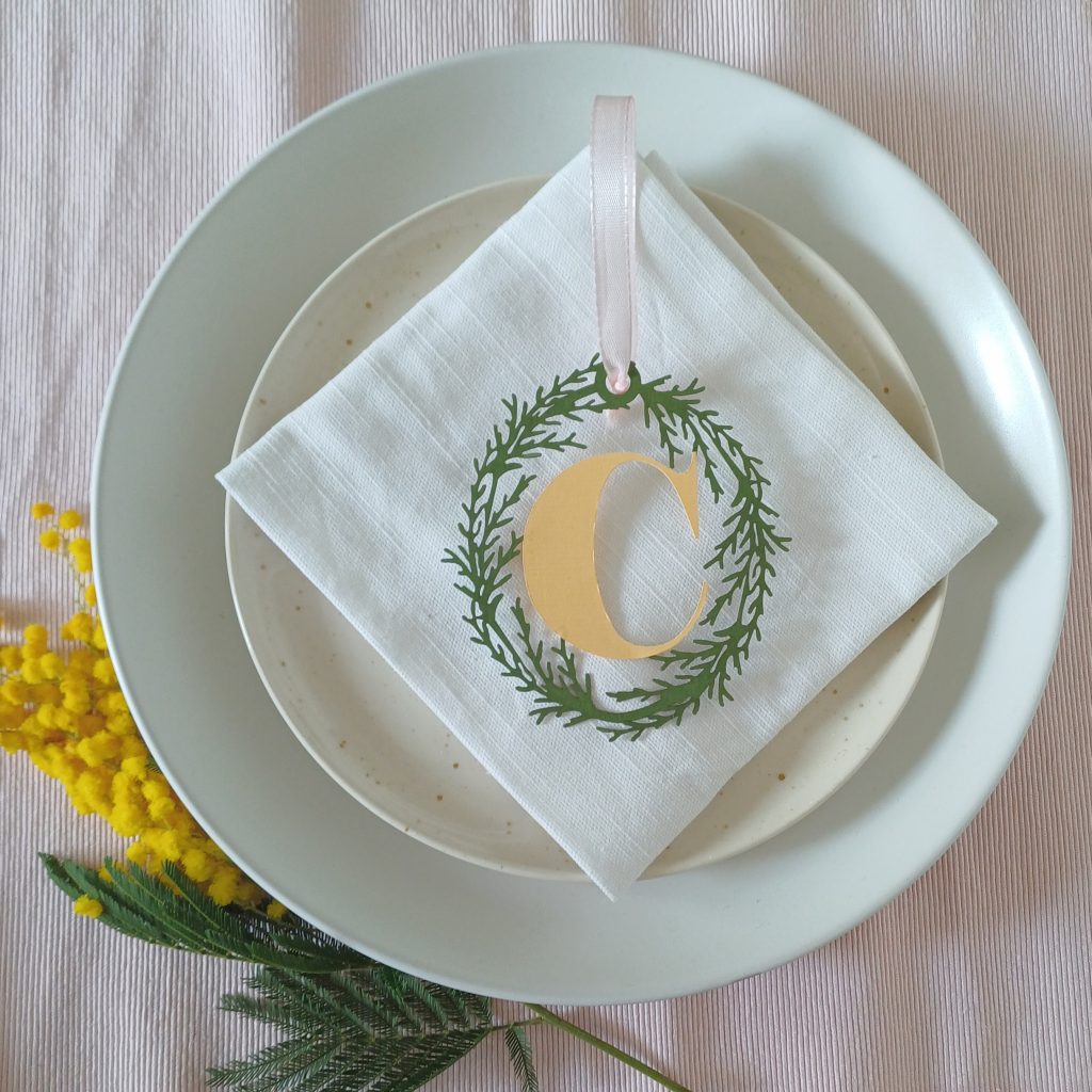 Sprig of mimosa tucked under two stacked white plates. A white napkin is in the centre of the plates with a papercut initial decoration placed on top.