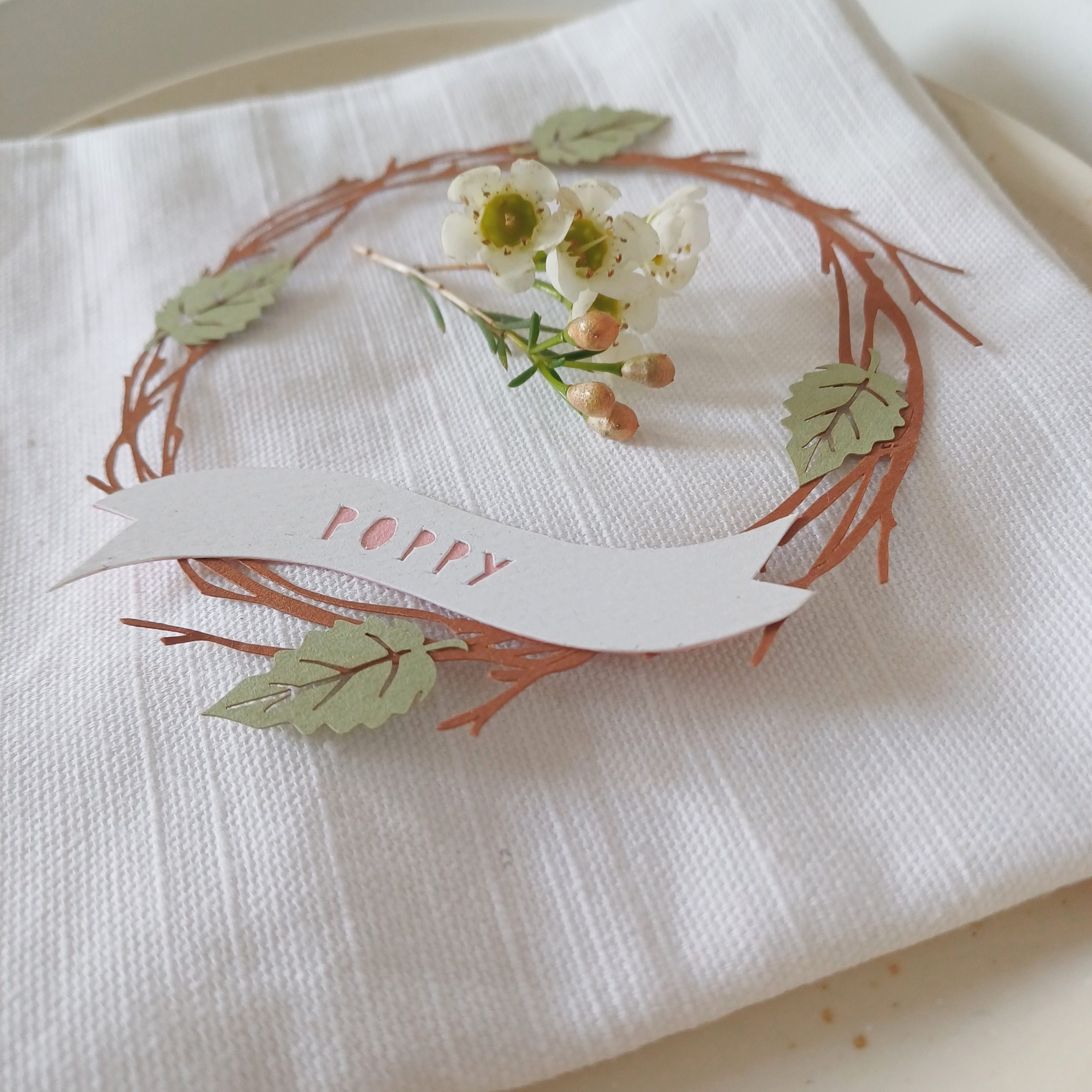 Close view of wreath papercut place setting placed on white napkin. In the centre of the wreath on top of the napkin is a sprig of wasflower blossom. Wreath is made from brown paper with 4 pale green leaves distributed at north, south, east and west positions. A white paper banner runs across the bottom of the wreath with letters cut out to reveal the name "poppy" in pink.