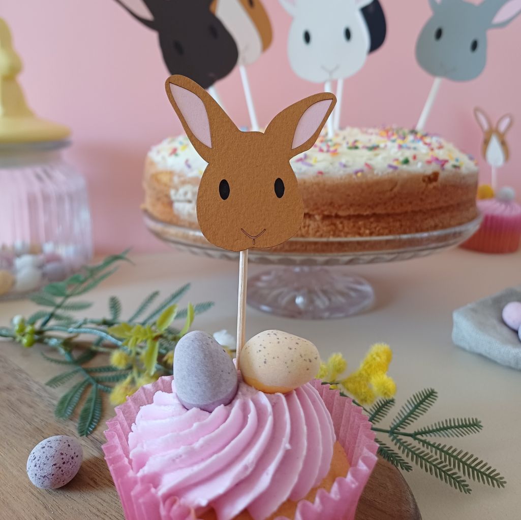 Closeup view of toffee brown easter bunny cupcake topper placed in a cupcake with pink icing. In background large sponge cake decorated with a corrdinating set of larger Easter Bunny Cake Toppers is visible